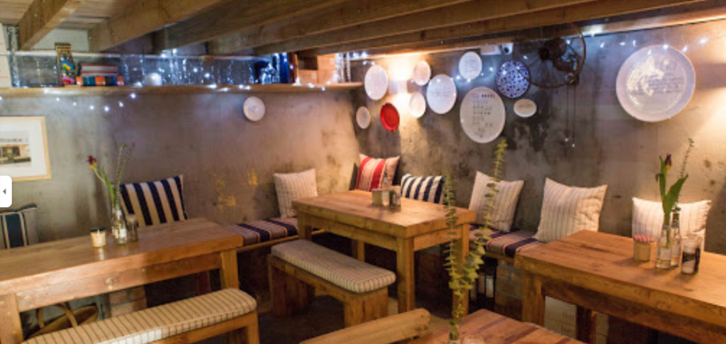 Cosy space with bare wood tables and benches, concrete walls surround it, fairy lights and decorations make it a lovely venue for breakfast Cape Town has to offer