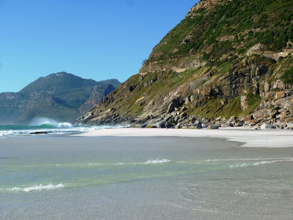 flat sandy beach with the surf coming over it, mountains loom over this Noordhoek beach one of many great Cape Town beaches