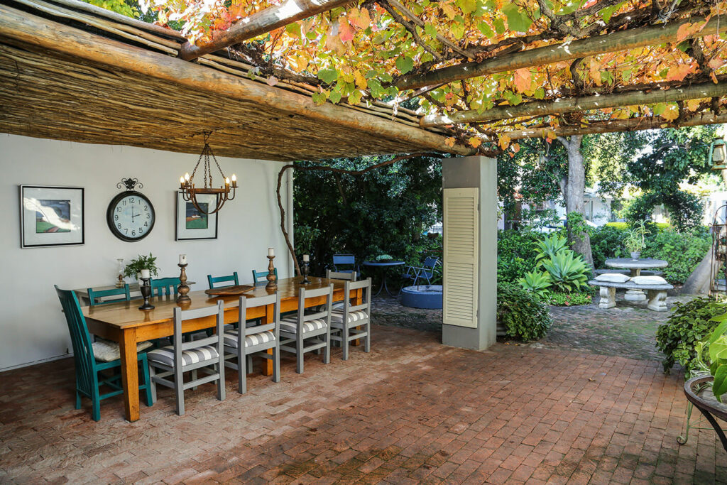 The patio at this Paarl accommodation, features a large table with 10 chairs under cover, and a more natural leafy cover as well, with tables visible in the garden as well