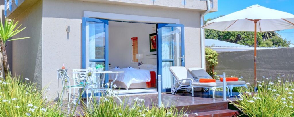 Loungers and table on a deck with open glass doors, through the doors you can see the luxurious room with bed, another great accommodation at Mossel Bay