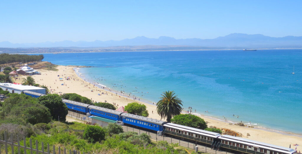 Train cars lined up just above a beautiful white sandy beach, the other side of the bay is visible on the horizon, a unique accommodation at Mossel Bay