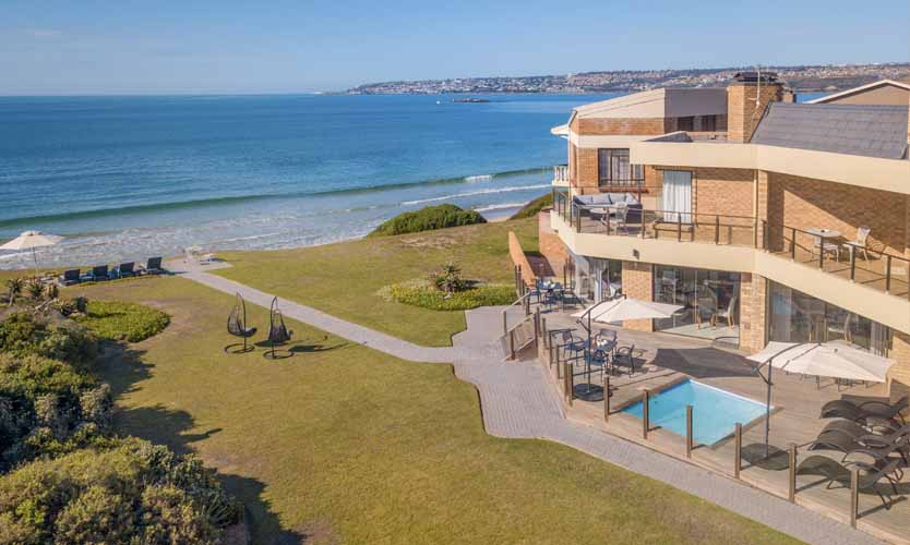 Plain brick double storey building with a small pool and lounging area located in front of it, grass stretches from the building to the beach, another fantastic accommodation at Mossel Bay