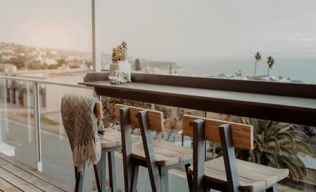 a rustic styled balcony with wooden stools lined up by the counter attached to the balcony barrier, in the background you can see the view of the ocean and town seen from this accommodation at Mossel Bay