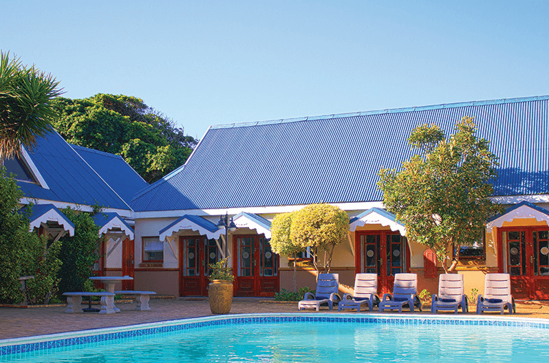 Beautiful swimming pool with sun loungers lined up next to it, the main building of the hotel with metal roofing stands behind it, a great pool at a fantastic accommodation at Mossel Bay