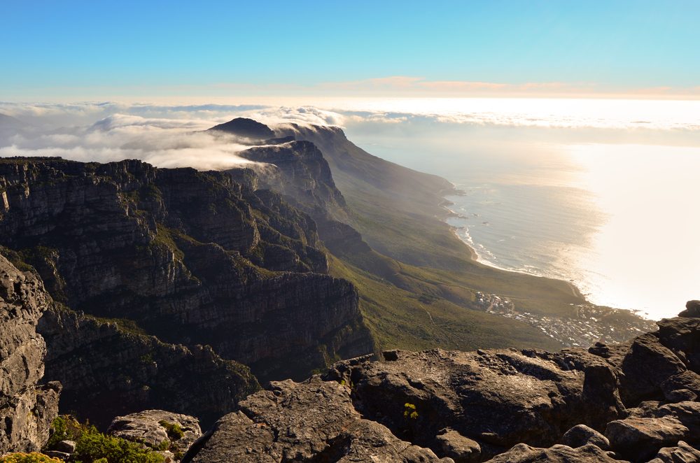 View from the top of Table Mountain, you are able to see the mountain range extending parallel to the coast, with clouds seemingly flowing over the cliff, this is easily the best SAN park in the Western Cape