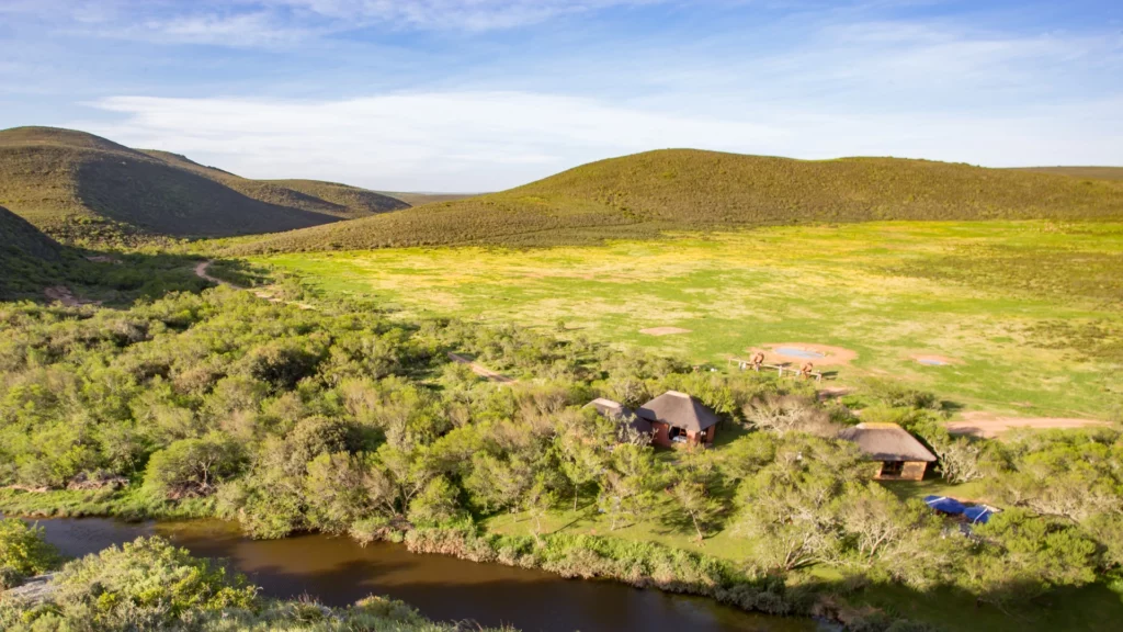 a river with a thick brush around it, a few thatch roof buildings can be seen amongst the brush, and behind them are wide open areas with hills in the distance, a great accommodation at Mossel Bay