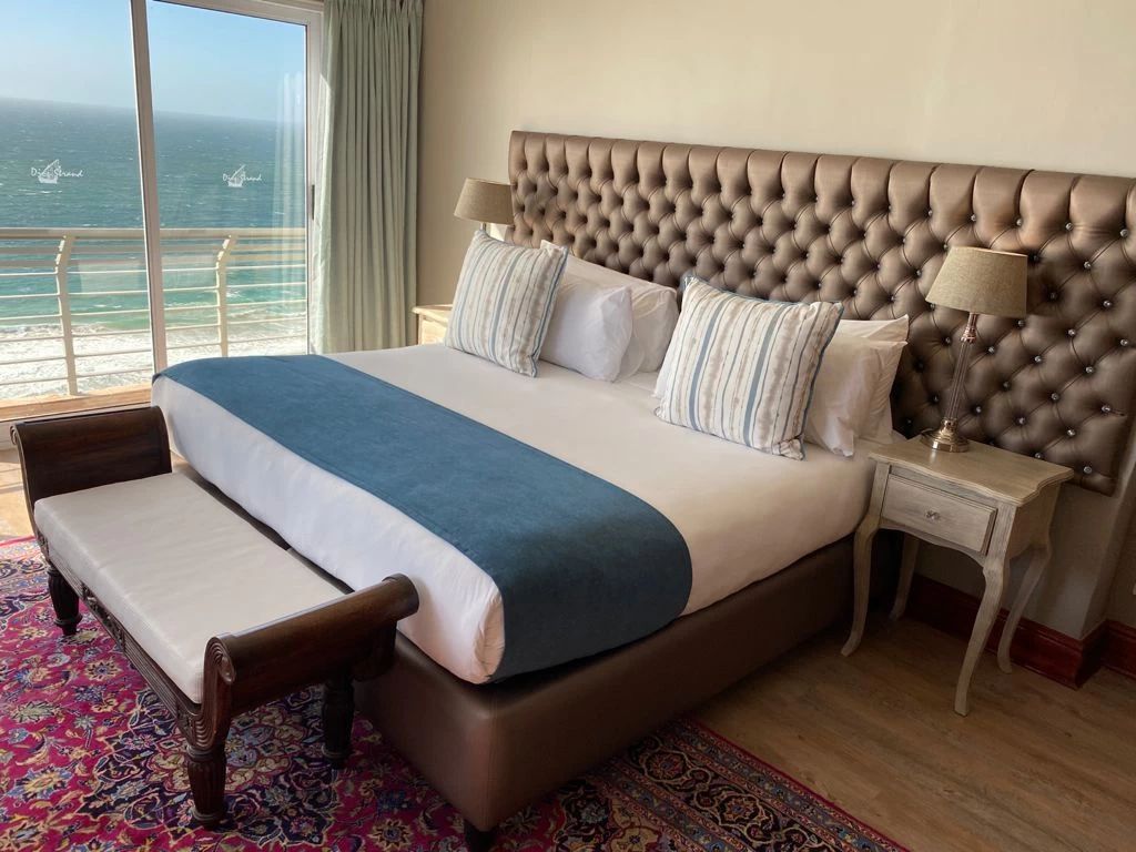 A large bed with an ottoman at the foot, a luxury headboard, glass doors lead onto a balcony overlooking the ocean, just one example of what accommodation at Mossel bay has to offer.