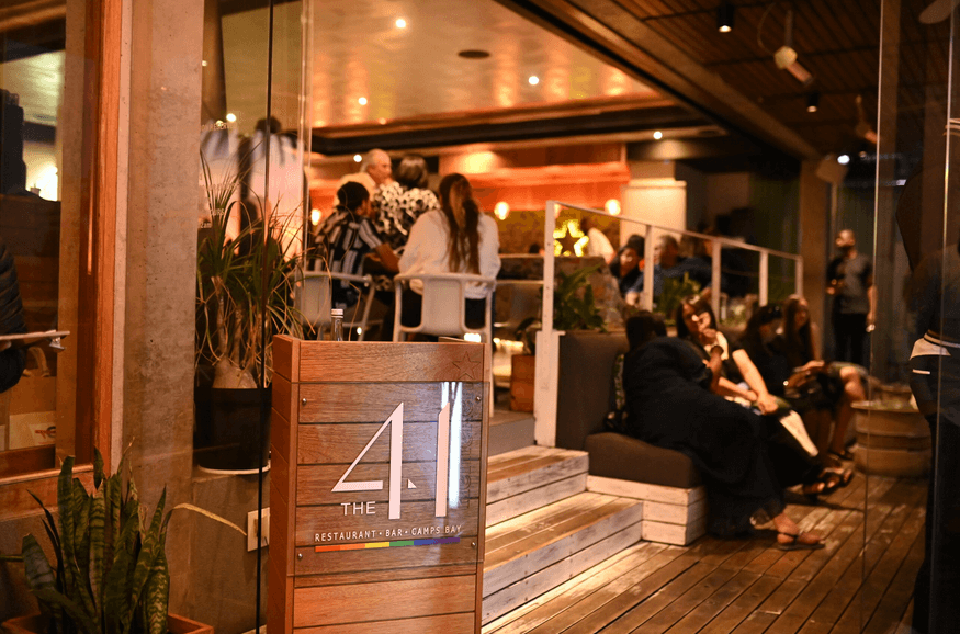 The front entrance of this restaurant in Camps Bay, with a wooden deck and well lit inviting interior